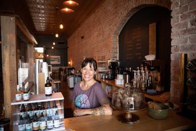Artisan Mark owner Elayne Woods Jones stands behind the coffeeshop's counter. Behind her, a long, exposed brick wall extends to seating at the back of the shop, with warm lights overhead.
